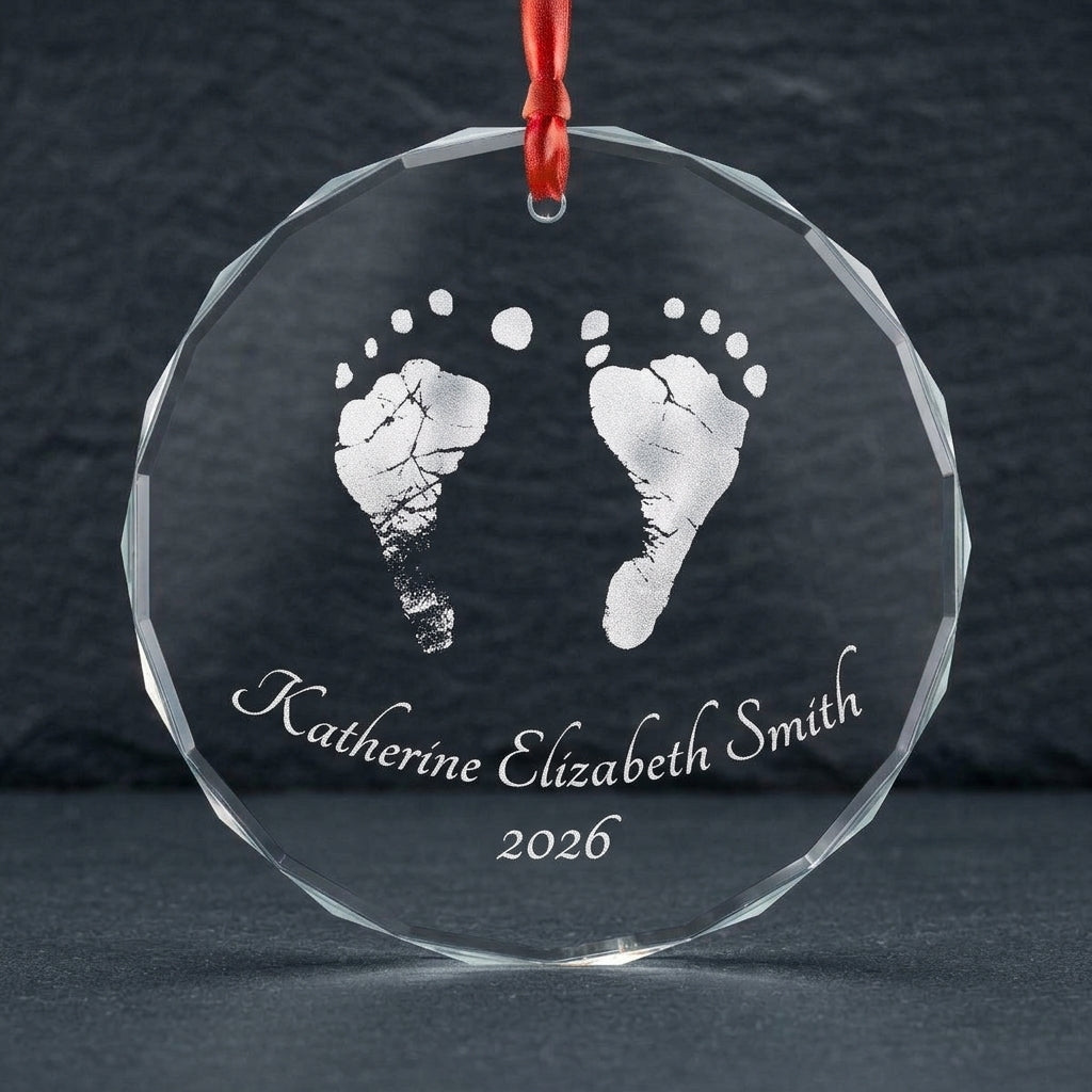 A personalized clear glass or acrylic round hanging ornament with a red ribbon loop, featuring engraved baby silver footprints and the name 'Katherine Elizabeth Smith' with '2026' on a dark gray background.