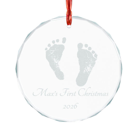 A round clear glass Christmas ornament with a red hanging ribbon, featuring a gray printed baby footprint and the personalized text 'Max's First Christmas 2026' on a white background.