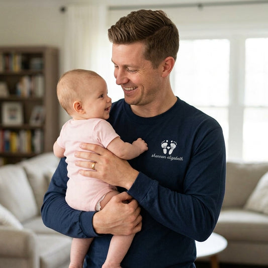 A man in a navy long-sleeve shirt holding a baby in a pink outfit, with a white custom print on the chest reading 'shannon elizabeth' with her baby footprints. The photo is taken in a bright, modern living room.