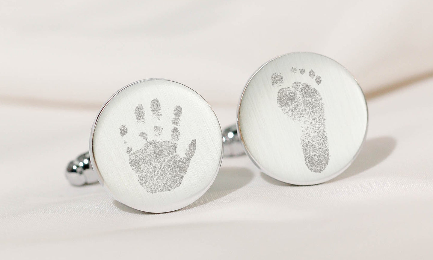 A pair of round silver keepsake cufflinks displayed on a soft beige fabric background. Each cufflink has an engraved baby print: a handprint on one and a footprint on the other.