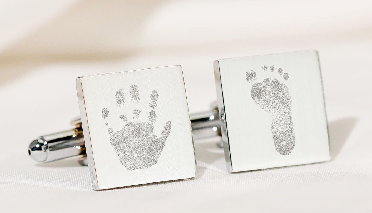 A pair of silver square cufflinks displayed on a soft cream fabric background, each engraved with a baby print (one handprint and one footprint). 