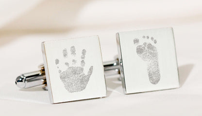 A pair of silver square cufflinks displayed on a soft cream fabric background, each engraved with a baby print (one handprint and one footprint). 