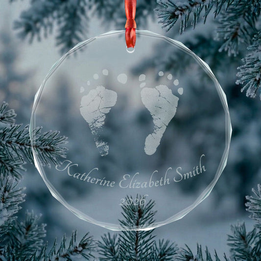 A personalized clear glass Christmas ornament keepsake hanging from a red ribbon, featuring engraved baby footprints and the name in script, set against a blurred winter pine background.