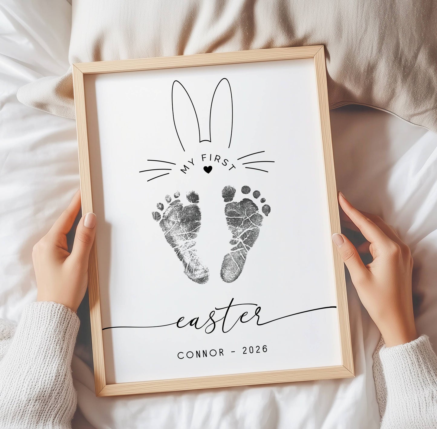 Framed print with footprints and text 'My First Easter Connor 2026' held by hands on a soft surface.