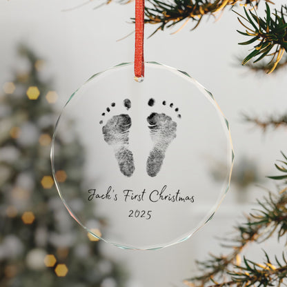 Custom glass ornament with footprints, First Christmas 2025' text, hanging on a tree branch.