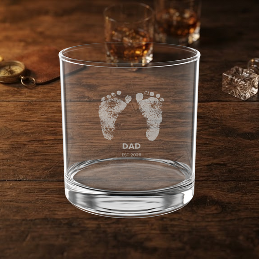 A clear glass rocks tumbler with etched baby footprints and the text 'DAD EST 2026' on a dark wooden table, with blurred whiskey glasses in the background.