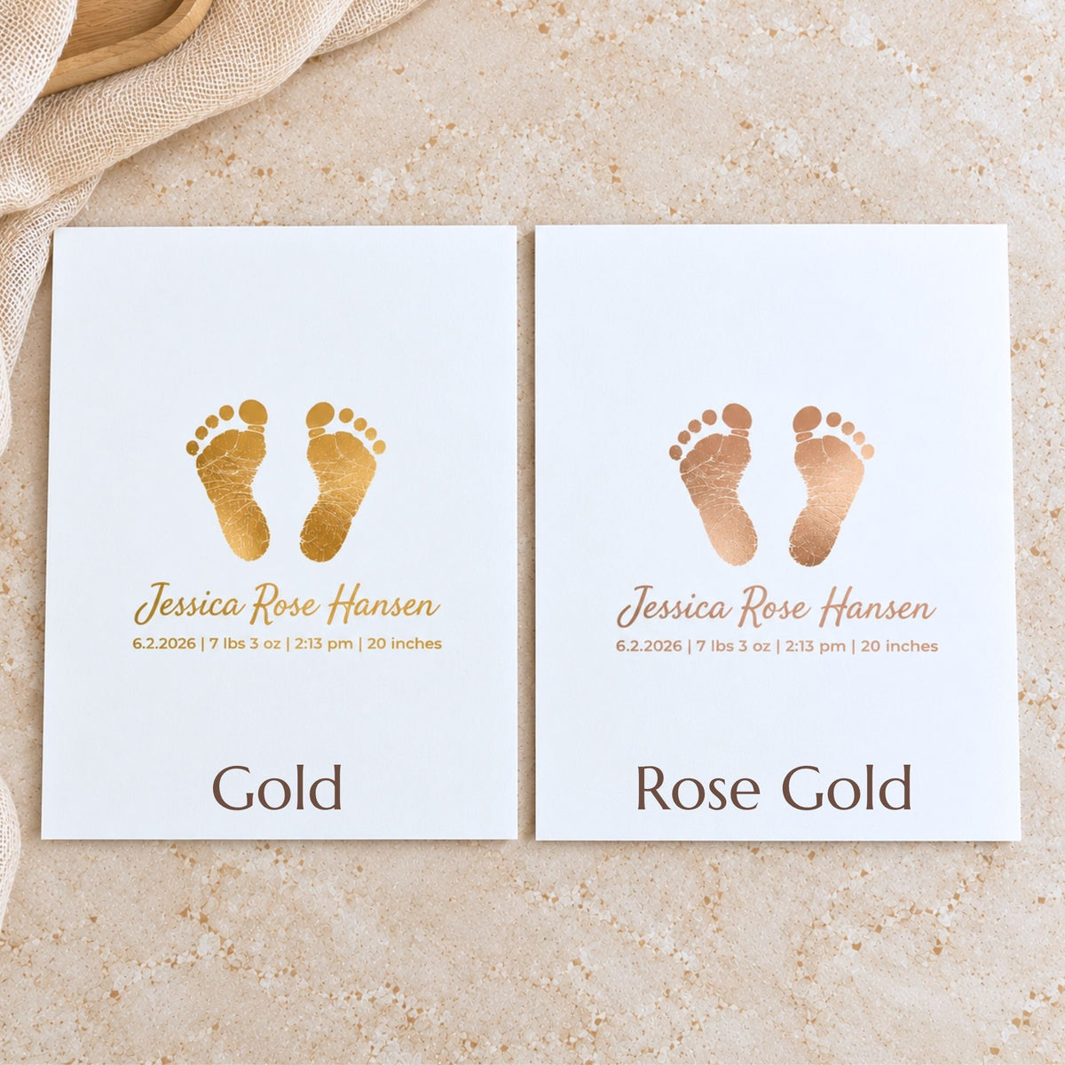 Two white baby keepsake cards with gold foil footprints and personalized names, displayed side by side on a textured beige stone surface with a beige fabric corner.