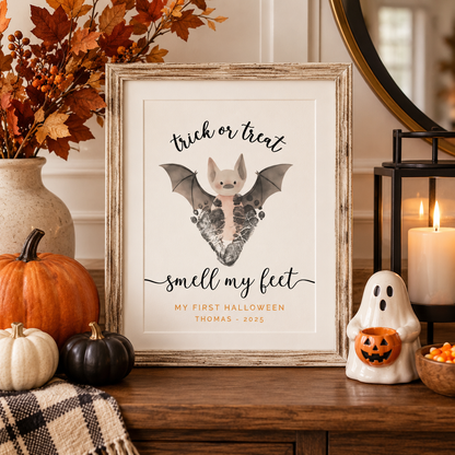 A framed personalized Halloween footprint keepsake featuring a cute bat baby on a heart-shaped boot with the text 'trick or treat smell my feet MY FIRST HALLOWEEN THOMAS - 2025'. The rustic wood frame is styled on a wooden table with autumn decorations including pumpkins, a ghost candle holder, and a lit candle.
