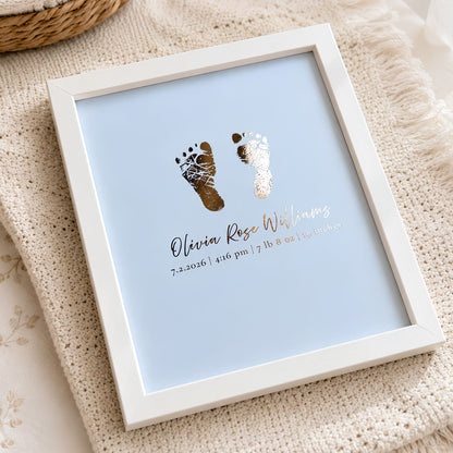A framed personalized baby keepsake print  keepsake featuring gold foil baby footprints and custom text on a light blue background, set in a white frame on a textured cream knit blanket.