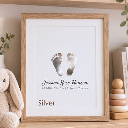 A personalized framed baby footprint print keepsake with silver foil footprints and the name 'Jessica Rose Hansen' on a white background, housed in a light oak wood frame and styled on a wooden shelf next to a plush bunny toy and books.