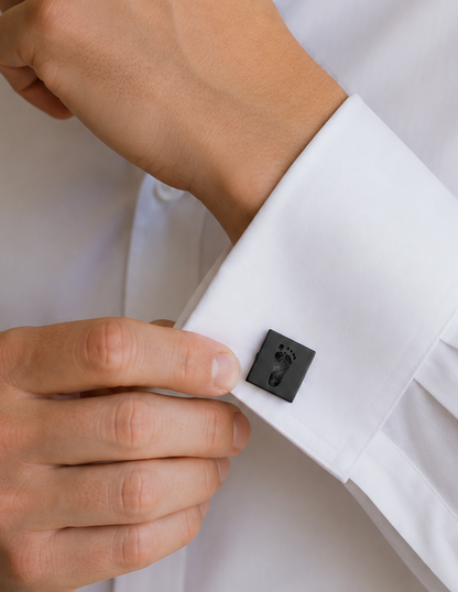 A close-up of a man wearing a white dress shirt and adjusting a black-toned keepsake cufflink with a baby footprint on it.