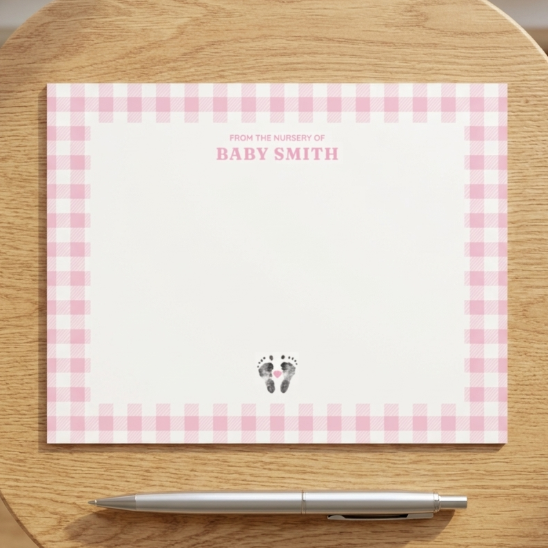 Stationery with pink checkered border and  baby footprints. 'Baby Smith' text on a wooden surface with a pen.