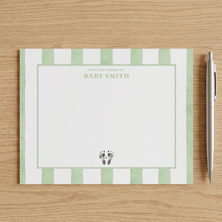 Note card with green stripes and baby footprint design on a wooden surface with a pen.