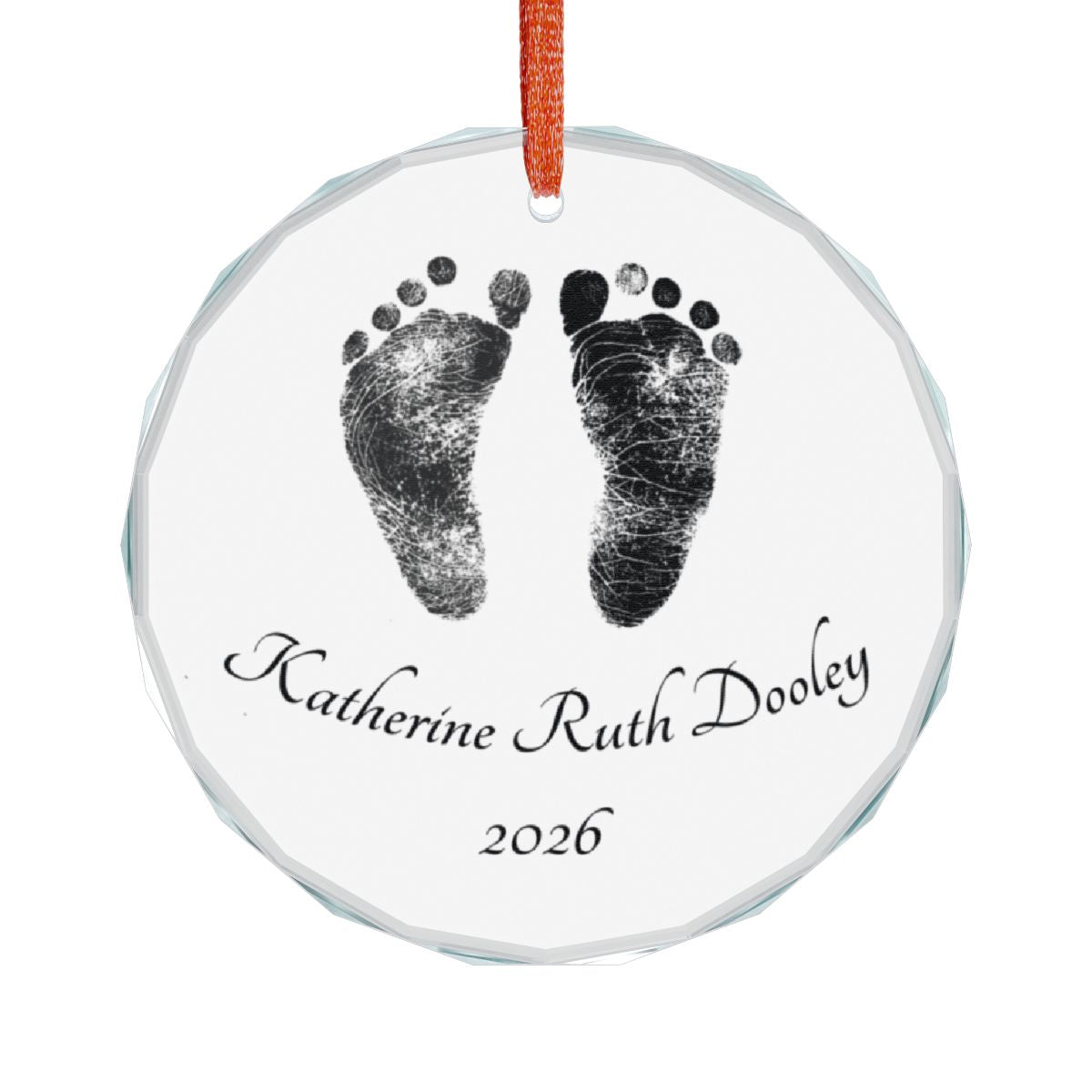 A round clear glass keepsake ornament featuring black footprints and the personalized name with the year '2026' printed below, hanging from an red ribbon on a white background.