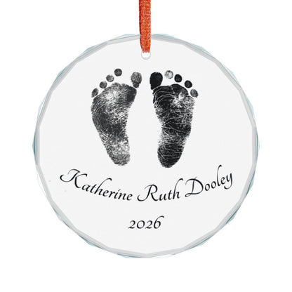 A round clear glass keepsake ornament featuring black footprints and the personalized name with the year '2026' printed below, hanging from an red ribbon on a white background.