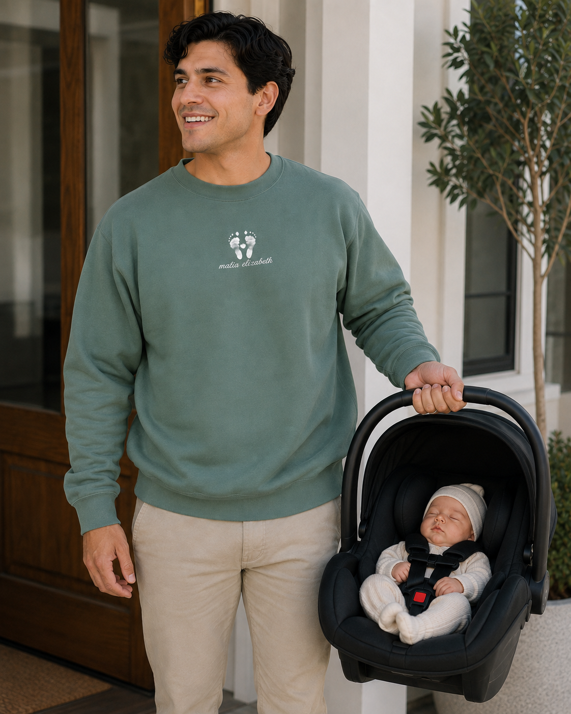 A man wearing a sage green crewneck sweatshirt with a white graphic logo of baby's footprints on the chest stands next to a baby in a black car seat, holding the baby seat handle in front of a modern house entrance.