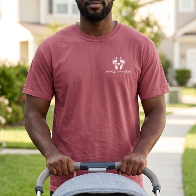 Man wearing a footprint t-shirt pushing a stroller with a baby inside on a sidewalk in a residential area.
